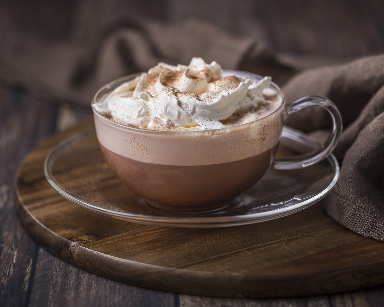 An ornate cup of hot chocolate with lots of cream, in a see through glass, sat on a wooden table.